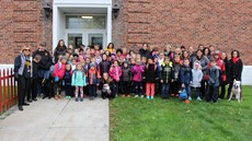 Group photo of dozens of elementary school students with parents and staff on the lawn in front of a brick building