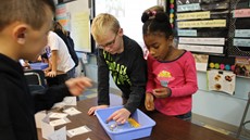 A young girl and boy look down at a blue bin on the desk in a classroom with another boy on the left side of the picture