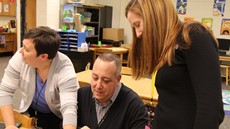 Lindsey Hendricks and Jenn Grasso stand to each side of a seated male teacher