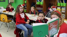 A female teacher wearing Santa ears holds a up a copy of The Grinch Who Stole Christmas while young students look on