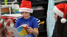 2nd-grade boy in a Santa hat holding a book while a high school female student sits next to him