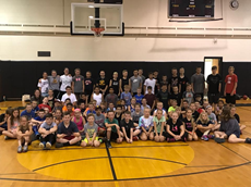 Group photo of dozens of children in t-shirts and shorts sitting on the high school gym floor with a row of bigger children standing behind them