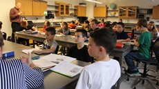 A classroom full of sixth-grade students look toward the front of the class while a teacher is standing in the left-hand side of the class