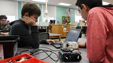 A small robot is on a table where two boys are sitting, looking at a laptop