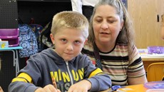 First grade boy looks at the camera as his grandmother sits behind him while he's at his desk at school