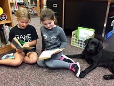 Two young girls sitting on a rug holding books, next to a black lab puppy