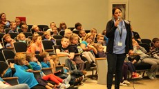 Woman holding a microphone with young students seated in an auditorium behind her