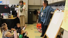 A teacher stands next to an easel with young students sitting in front of her