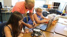 Teenage girl standing over a young girl at a desk with a laptop on it. 