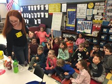 A woman standing as a class full of young students sits on a rug