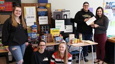 Five high school girls in a classroom standing or sitting near desks with boxes of food on them