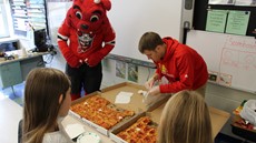 A man and dog mascot stand near two pizzas with two young girls