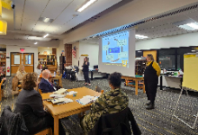 Two women speaking in front of a table of people