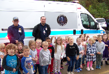 A group of small children standing wtih two men in front of an ambulance