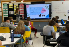 A woman standing in the front of an elementary classroom with children sitting at desks