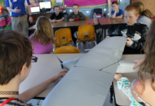 Four children sitting at a desk, holding a model airplane