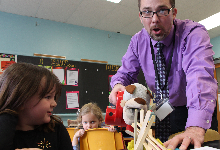 A man holds a hand-held vacuum in front of a pile of popsicle sticks as a young child looks on