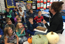 A group of young children sitting in front of a woman who has pumpkins to her side