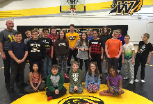 Over a dozen children standing on a wrestling mat smiling