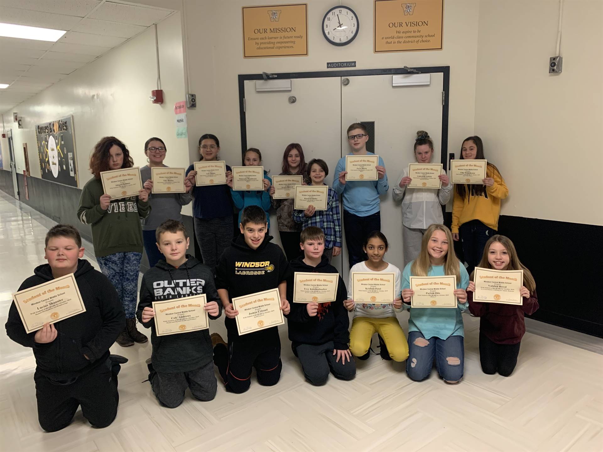 16 students holding certificates in a hallway