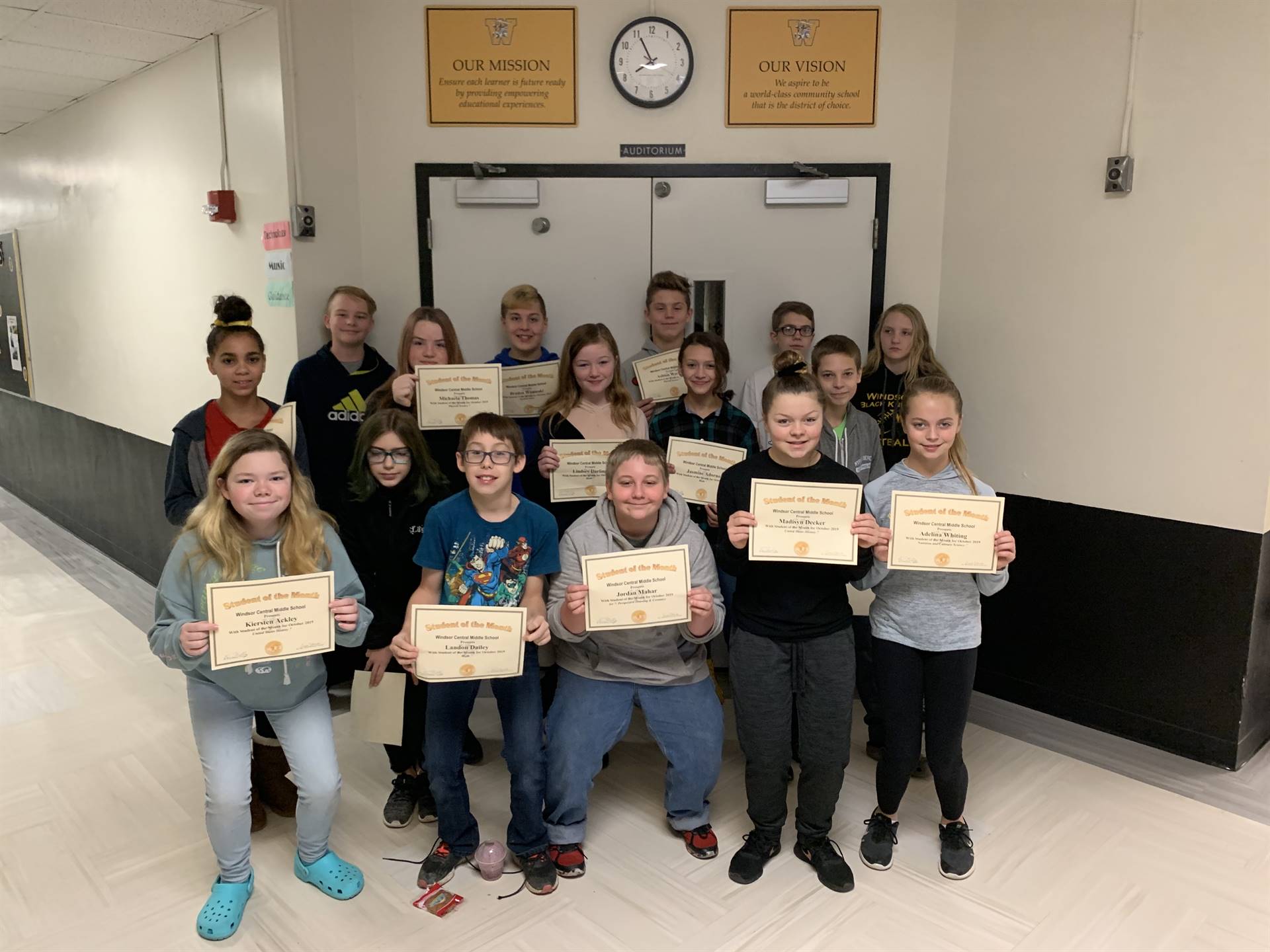 16 students holding certificates in a hallway